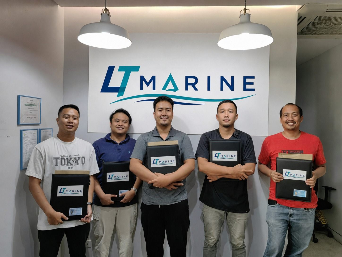 Five Filipino seafarers standing in front of the LT Marine logo wall, each holding a branded LT Marine document folder with their seafarer ID inside, during the final documentation handover before deployment.